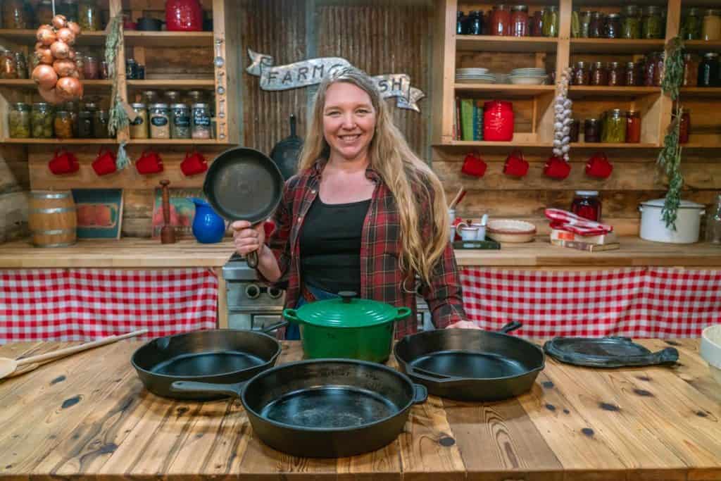 A woman holding a cast iron pan standing behind a table filled with cast iron cookware.