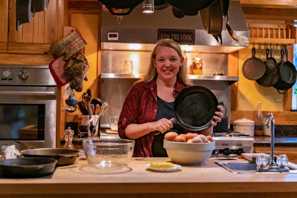 A woman holding up a cast iron pan behind a counter with eggs in a bowl.