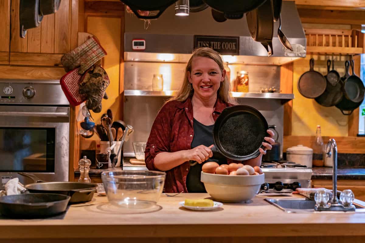 A woman holding up a cast iron pan behind a counter with eggs in a bowl.