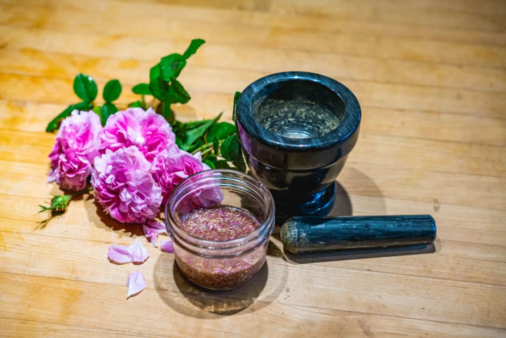 Vertical view of rose face wash in a mason jar next to fresh roses and a mortar and pestle.