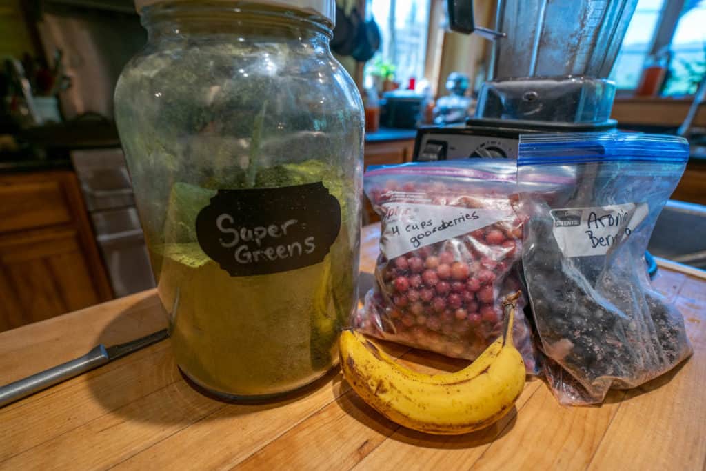 Super Greens Powder in a gallon jar with label next to frozen fruit and other smoothie ingredients.