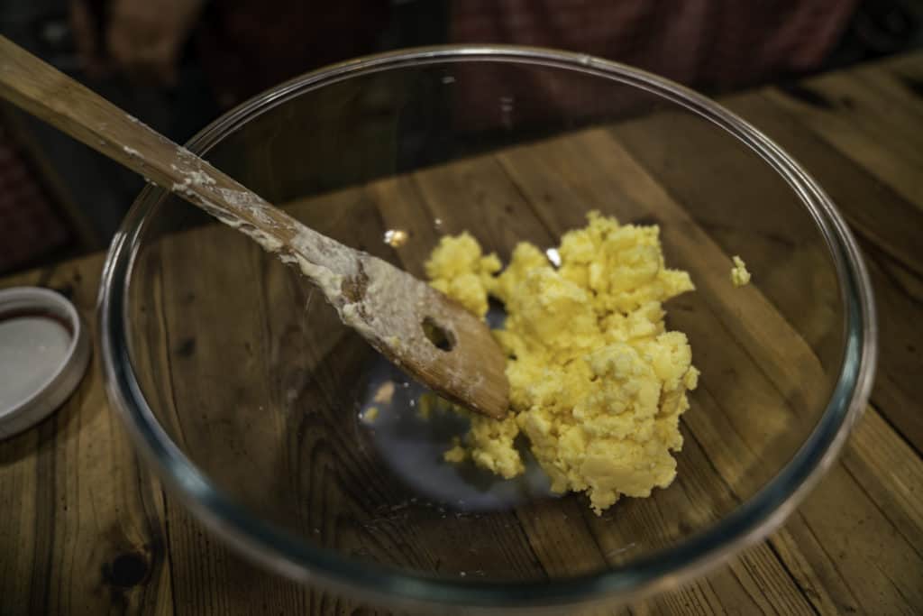 A clear glass bowl filled with butter and a wooden spoon pressing out the buttermilk.