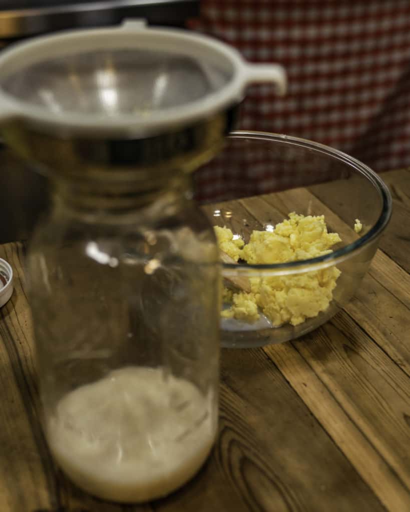 A quart sized jar with strainer and buttermilk inside with a bowl of butter in the background.