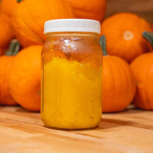 A jar of pumpkin puree sitting on a counter in front of a pile of pumpkins.