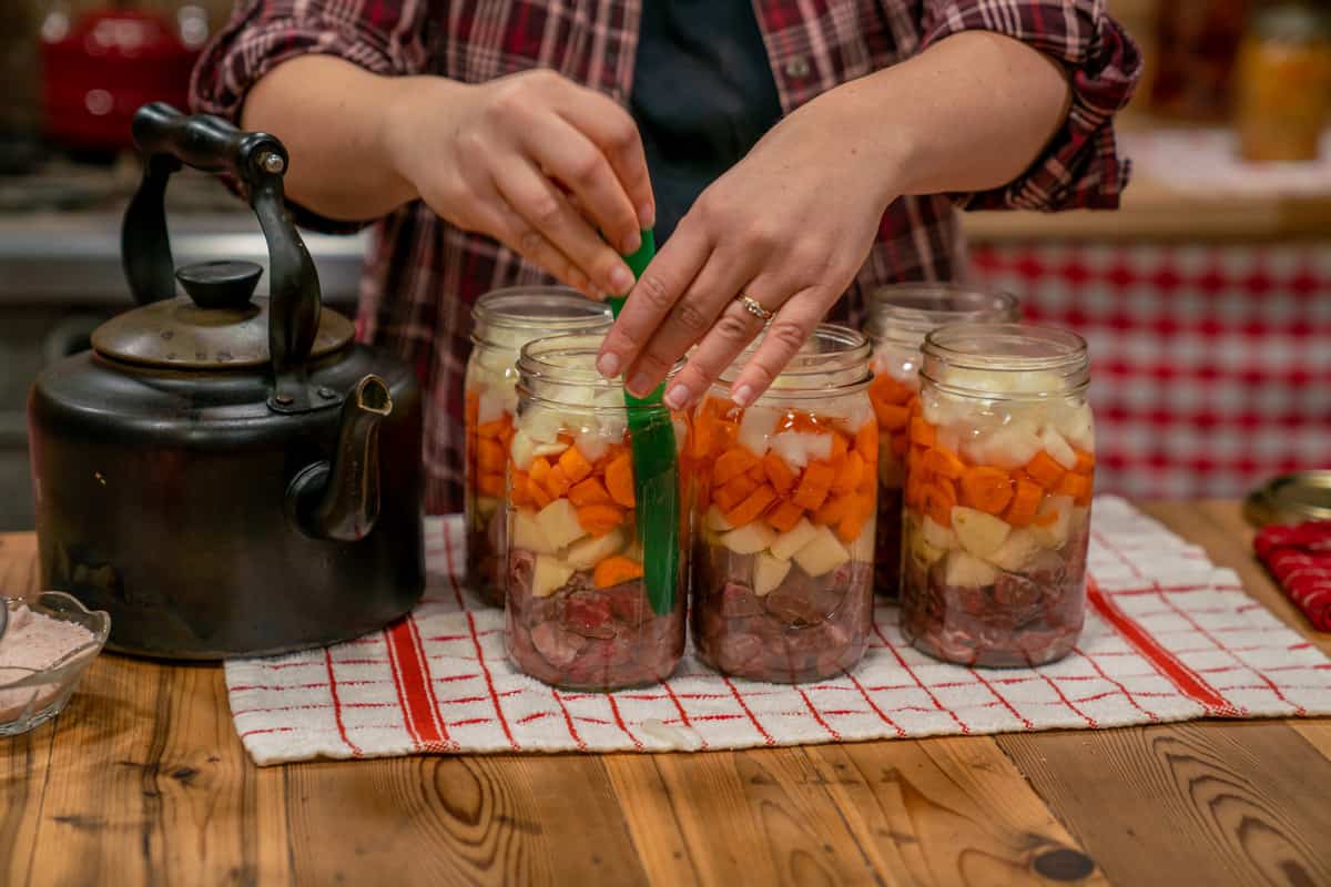 Six jars of raw packed canned stew with a canning tool sliding into one jar to remove air bubbles.