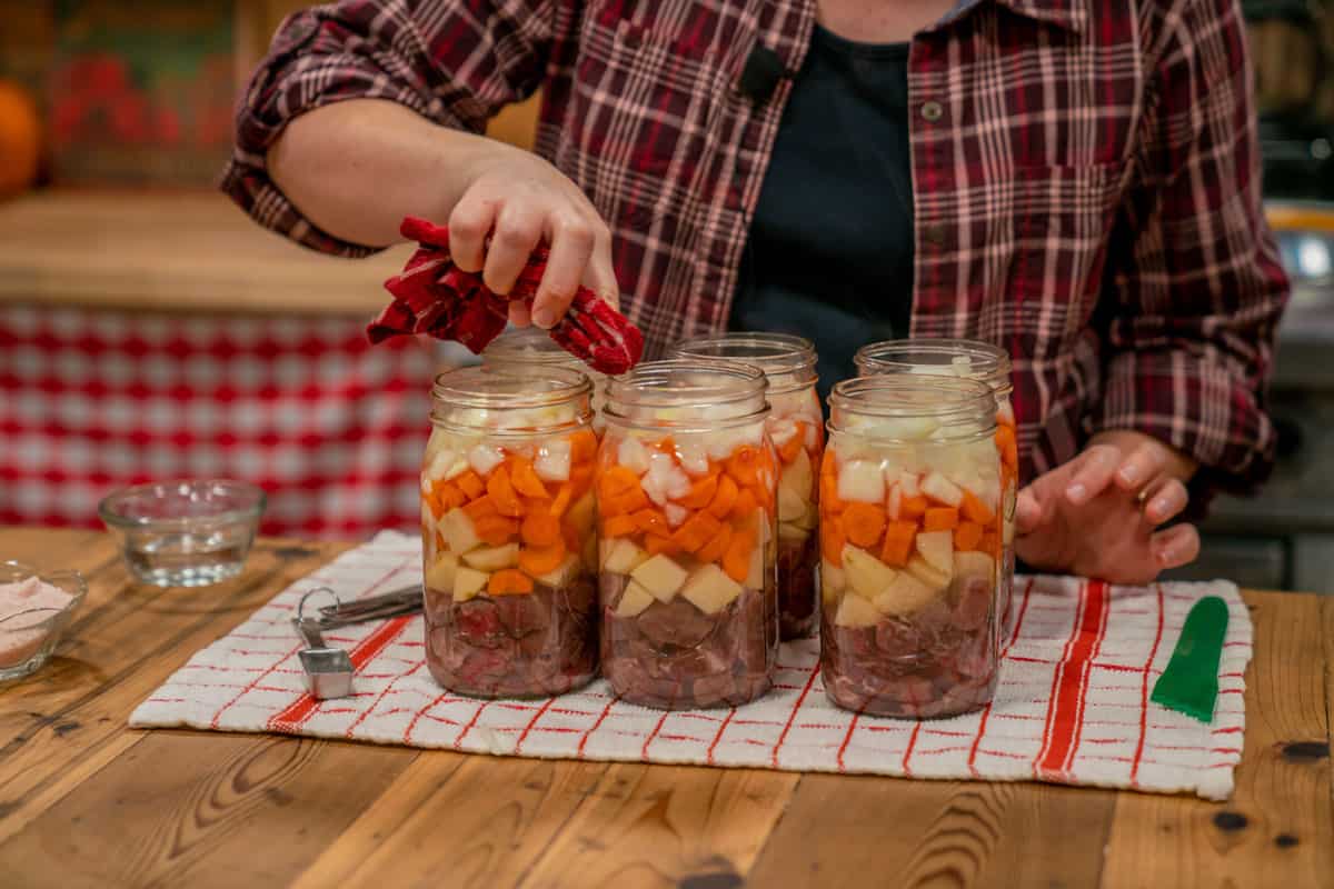 A woman wiping the rims of her canning jars filled with beef, potatoes, onions, and carrots.