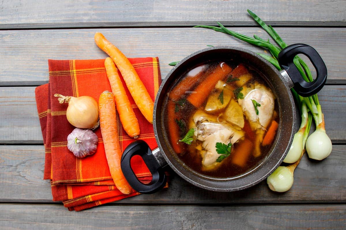 Chicken broth in a large pot with fresh veggies on the counter.
