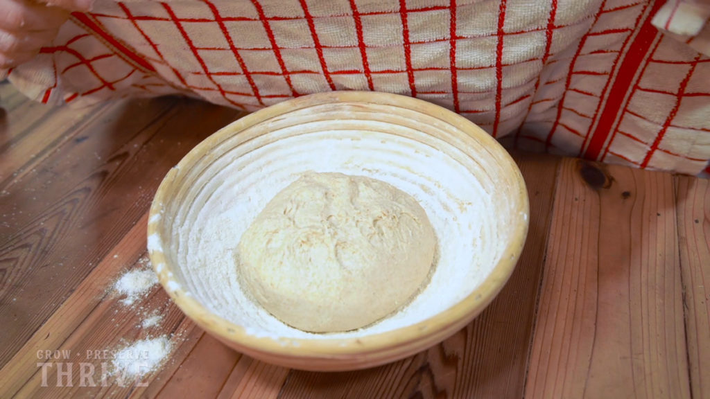 A ball of bread dough in a floured banneton being covered by a towel.
