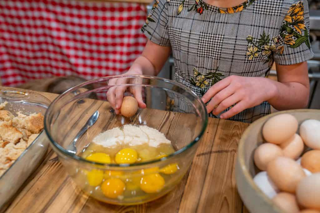 A young girl cracking an egg on the counter with a bowl filled with eggs in front of her.