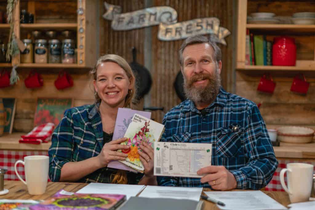 A man and woman sitting in the kitchen holding up a garden planner and garden planning books.