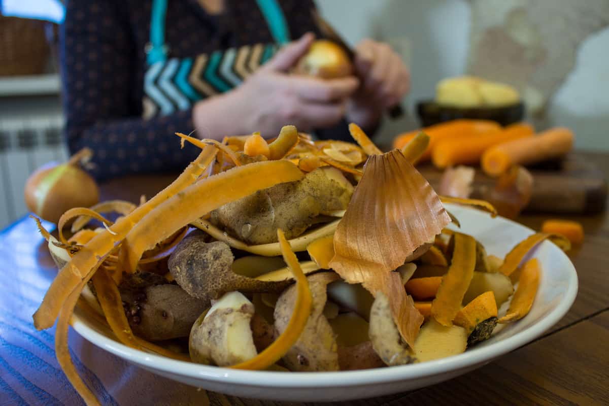 A plate of vegetable scraps on a table with a woman in the background peeling vegetables.
