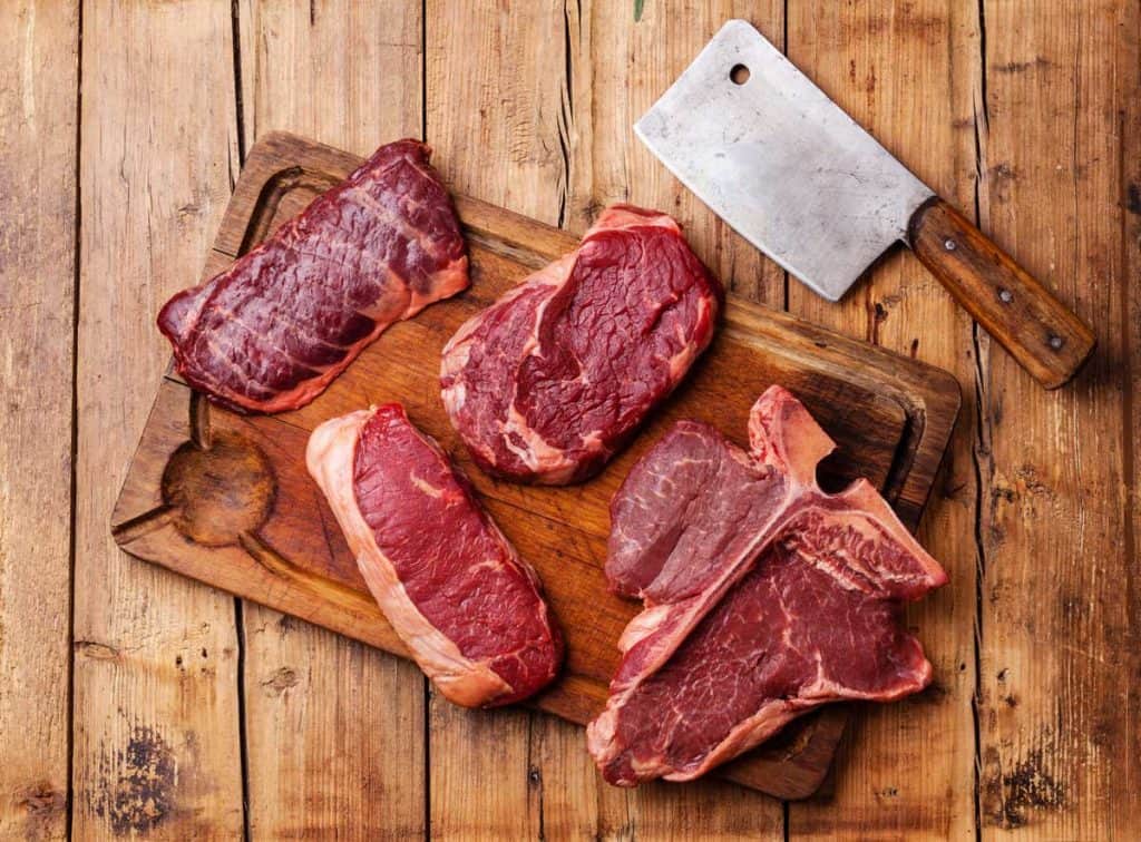 Various cuts of beef on a wooden cutting board with a butcher knife next to them.