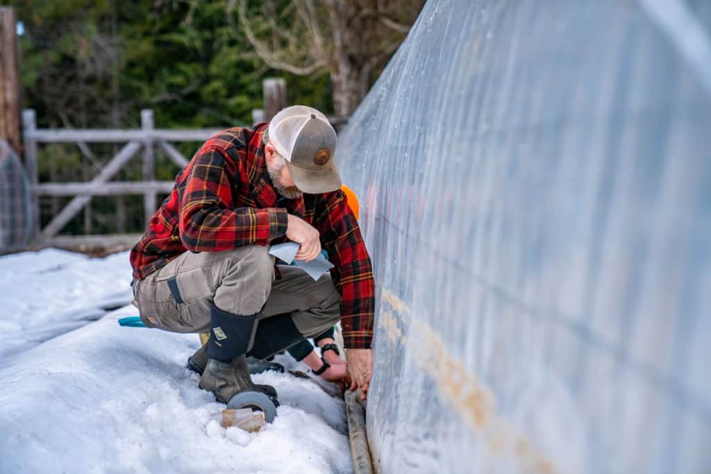 A man wrapping a hose with greenhouse plastic and securing it with gorilla tape.