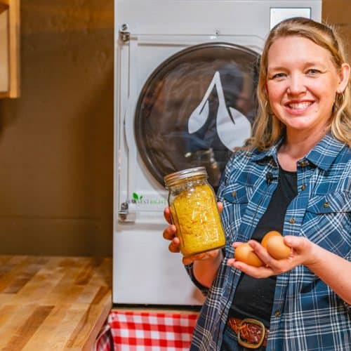 A woman standing in front of a harvest right freeze dryer holding a jar of powdered eggs in one hand and farm fresh eggs in the other hand.