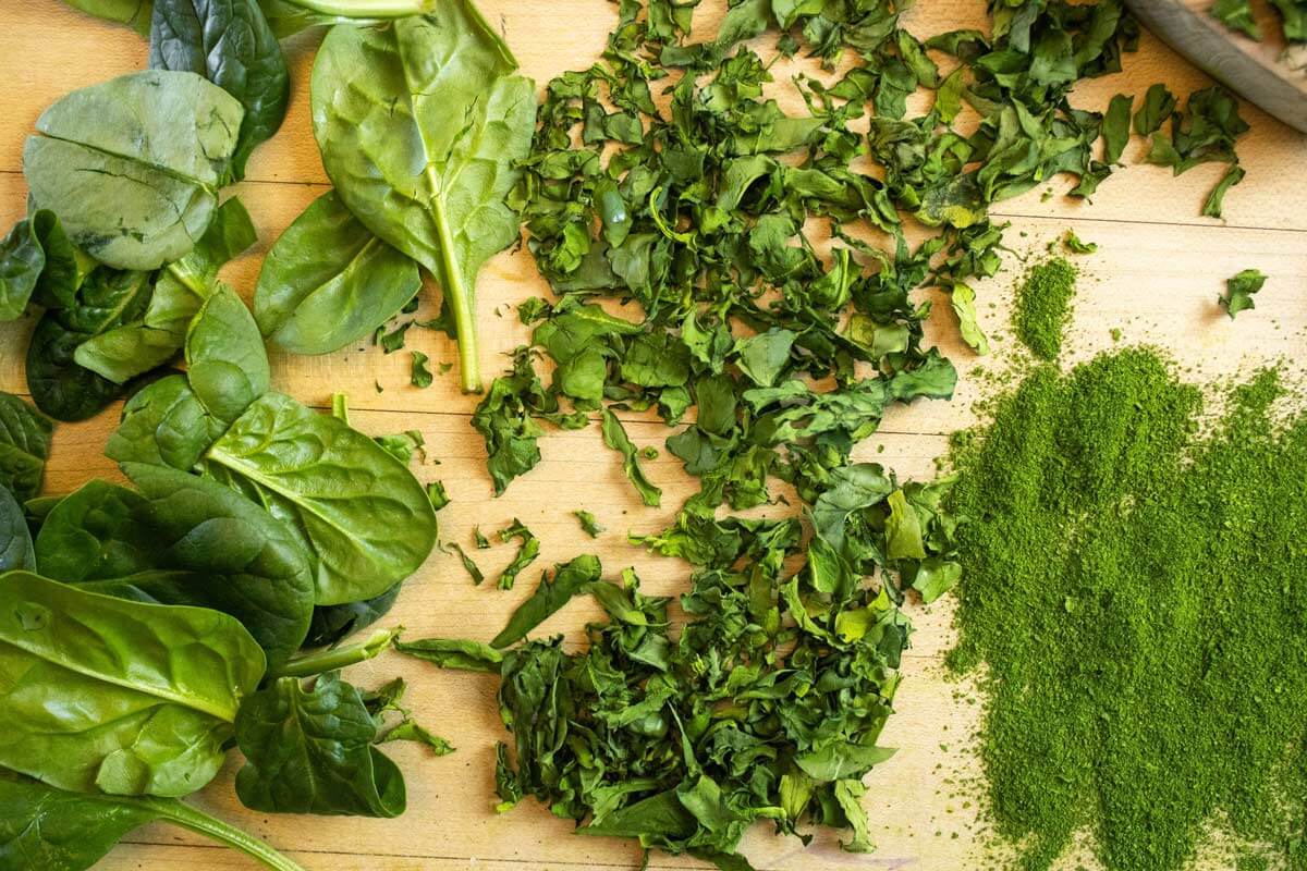 Whole leaves of spinach, chopped spinach, and spinach powder arranged on a wooden cutting board.