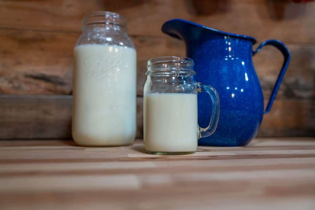 A pitcher, a half gallon jar, and a glass of milk on a counter.