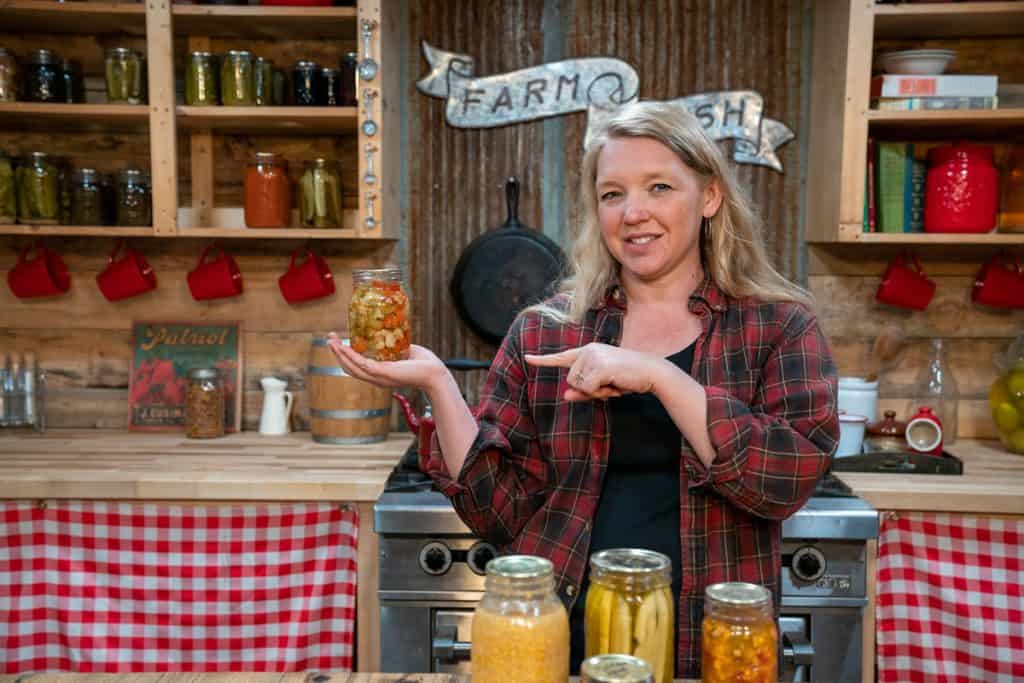 A woman holding up a jar of home canned food pointing to it. More canned food on the counter in front of her.