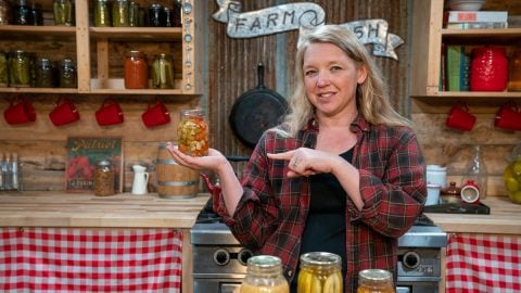 A woman holding up a jar of home canned food pointing to it. More canned food on the counter in front of her.
