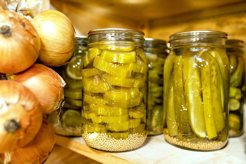 Canned pickles on a shelf with hanging onions to the side.