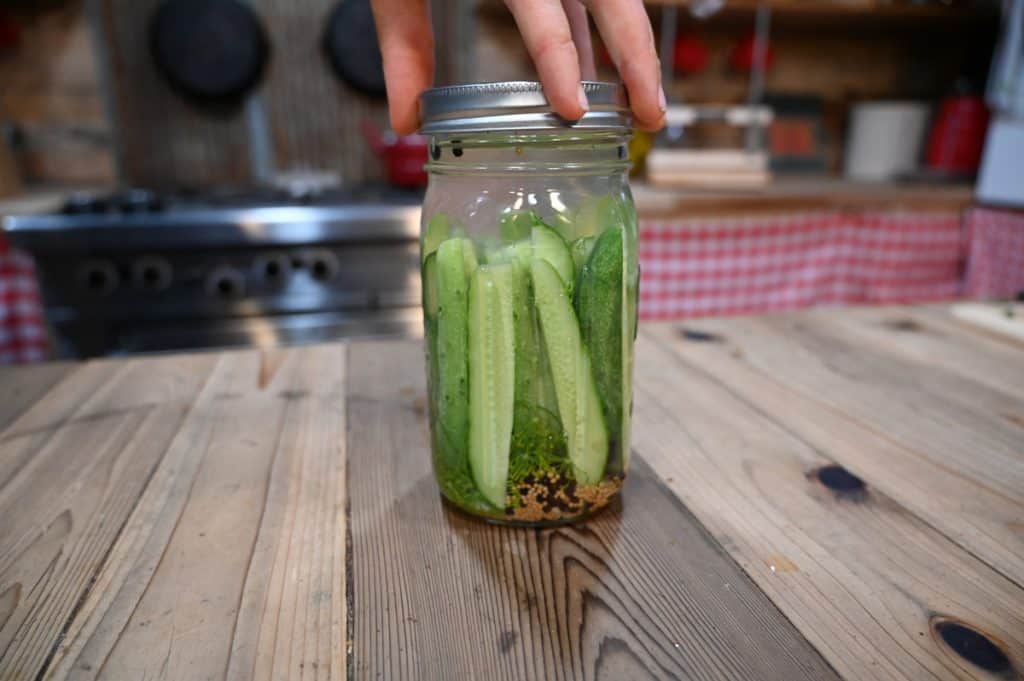 A hand screwing a lid onto a mason jar filled with pickle spears.