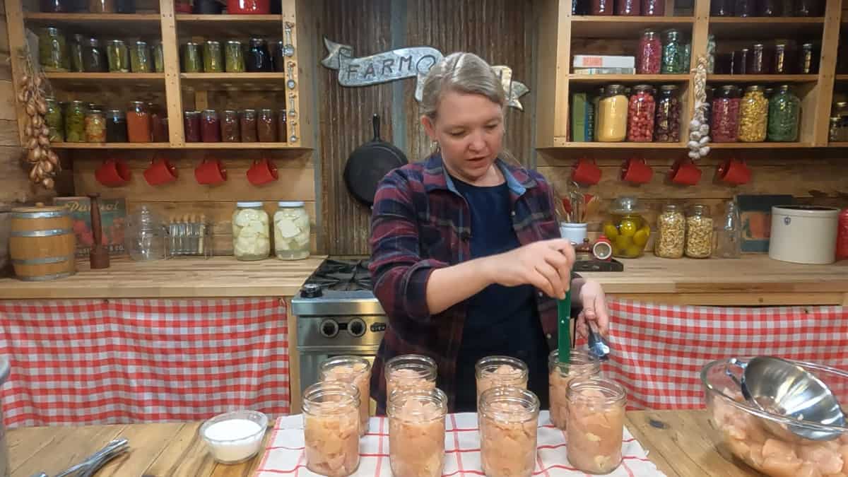 A woman using a bubble remover canning tool to press raw chicken into Mason jars.