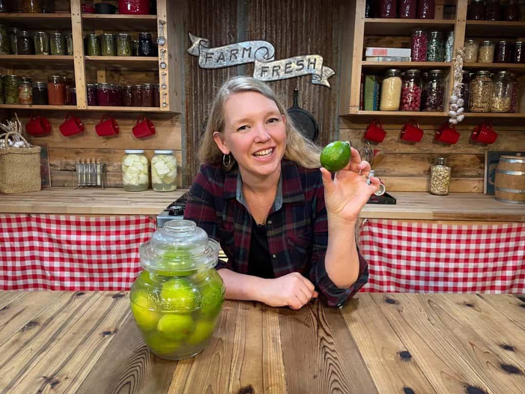 A woman smiling holding up a fresh lime next to a crock of fermenting limes.