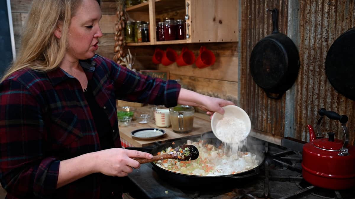 A woman sprinkling flour over a skillet of chicken pot pie filling.