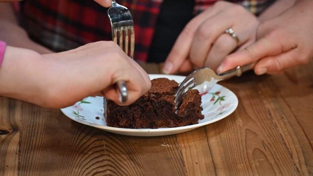 A brownie on a plate with multiple hands taking a bite.