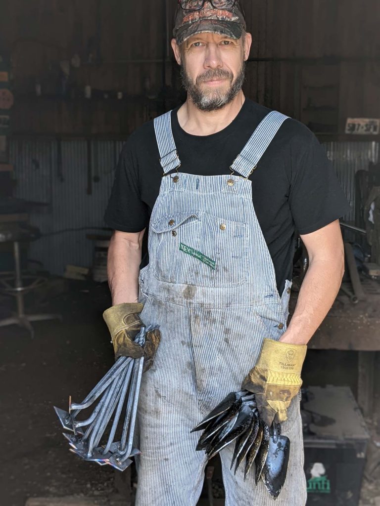 A man holding multiple hand-forged garden tools.
