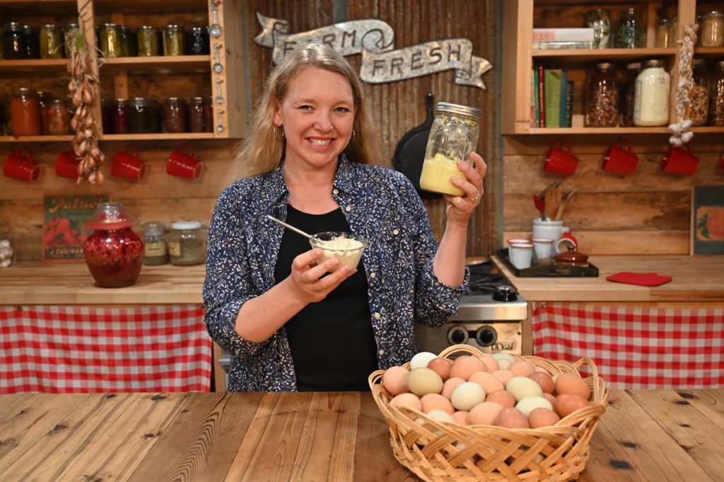 A woman holding a bowl and a jar of mayo with a basket of eggs on the table.