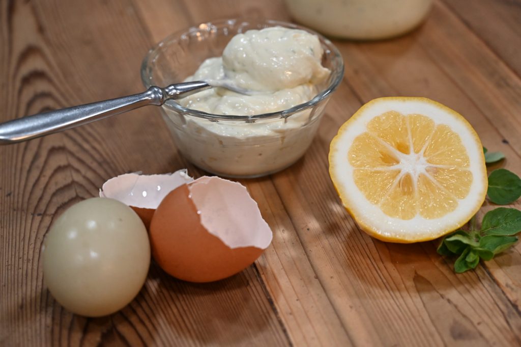 A bowl of mayo with a spoon, cracked eggs and a halved lemon on a counter.