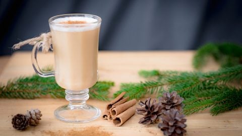 Homemade eggnog in a glass on a counter with pinecones and cinnamon sticks.