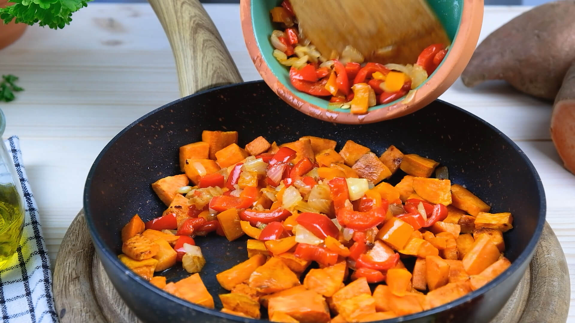 Pouring sauteed peppers and onions from a bowl into a cast iron skillet filled with cooked sweet potato.