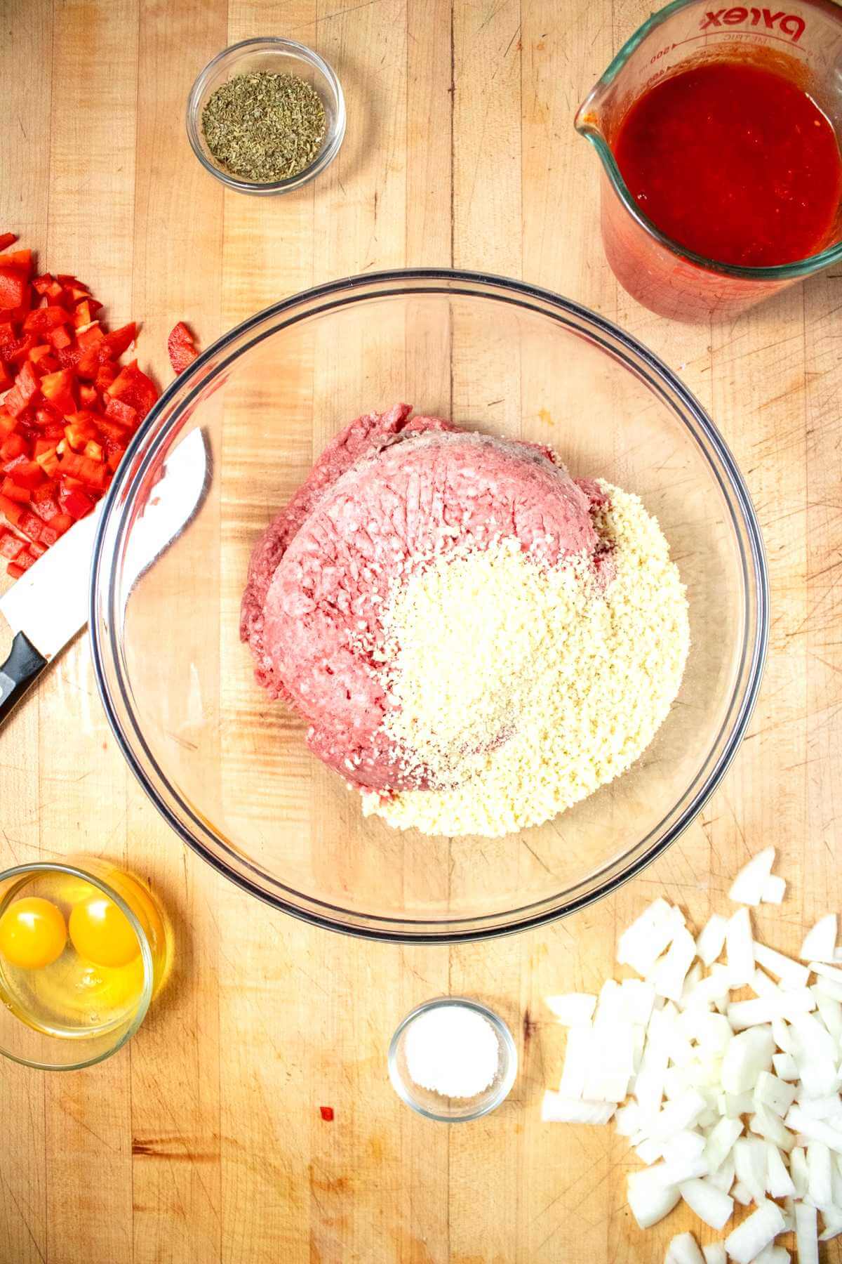 Ground beef and bread crumbs in a mixing bowl with prepared vegetables nearby.