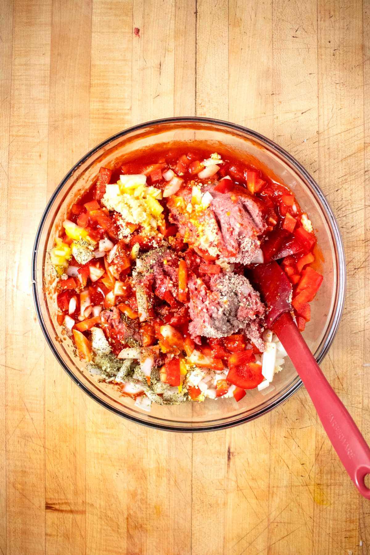 Ground beef, chopped vegetables and sauce being mixed in a glass bowl.