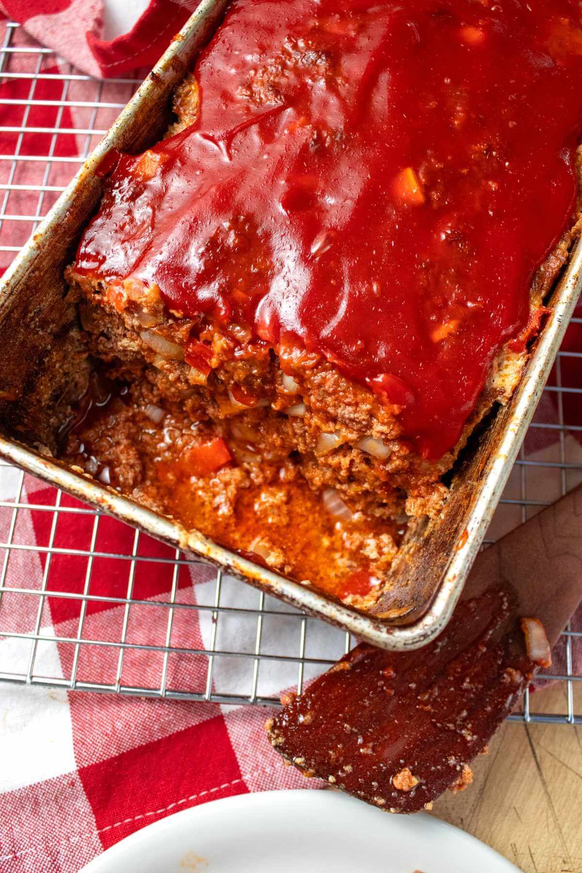 Glazed meatloaf in a loaf pan with a serving spatula beside it.