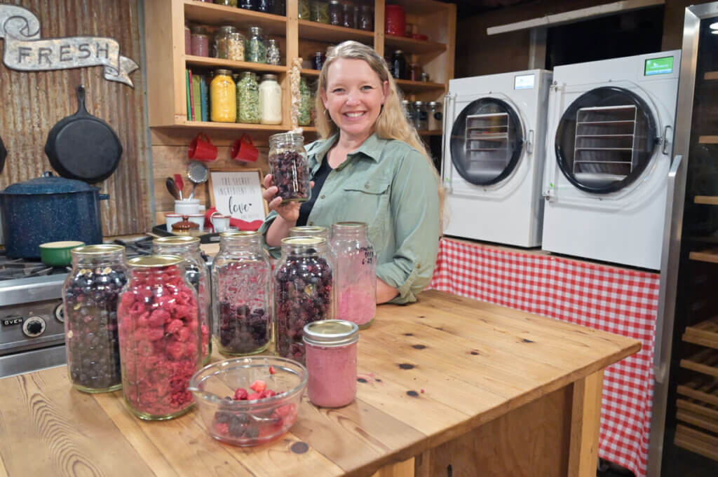 A woman standing behind jars of freeze dried fruit with two freeze dryers behind her.