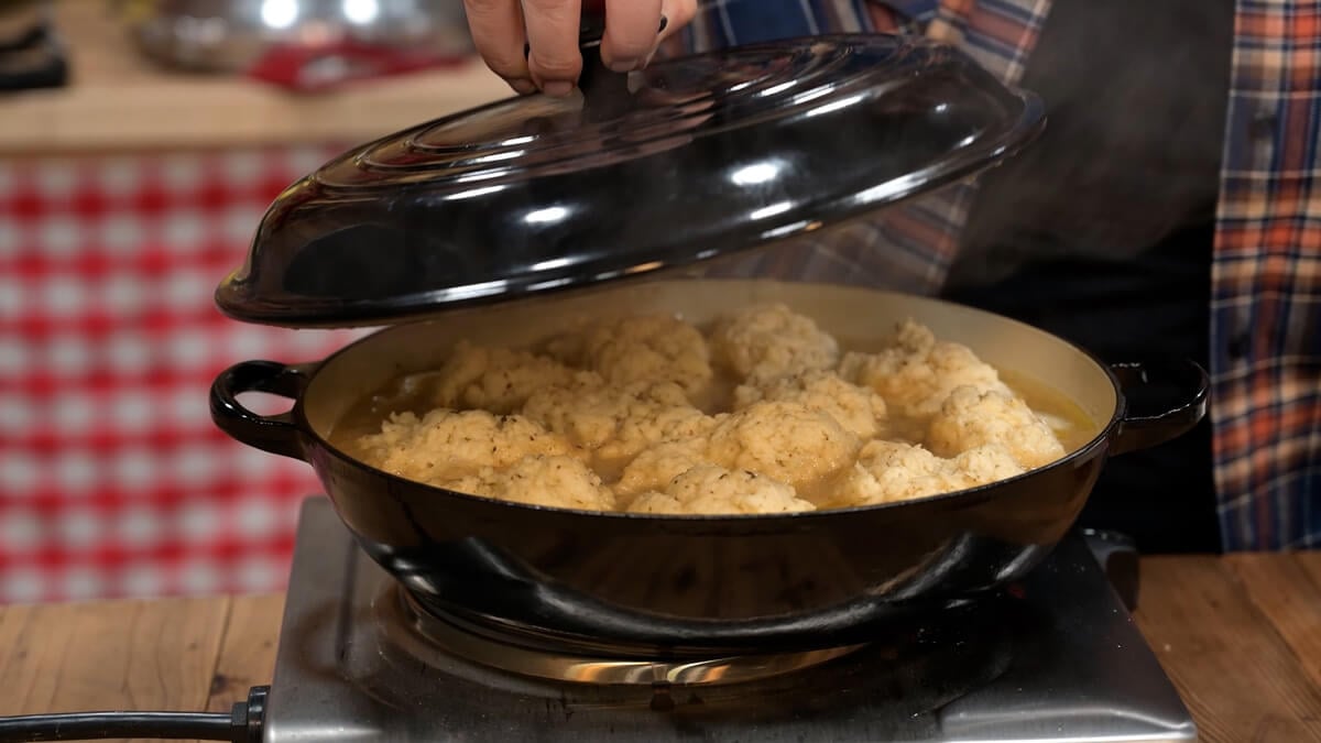 A woman lifting the lid off a pan of chicken and dumplings.