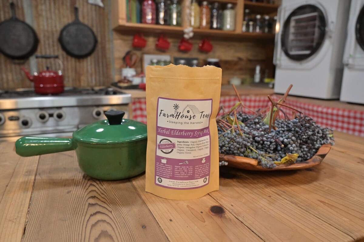 A pot, a basket of fresh elderberries and a pouch of elderberry syrup mix on a wooden counter.