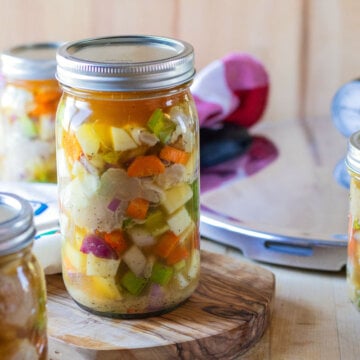 Lidded jars of chicken and vegetables on a wooden counter beside canning equipment.
