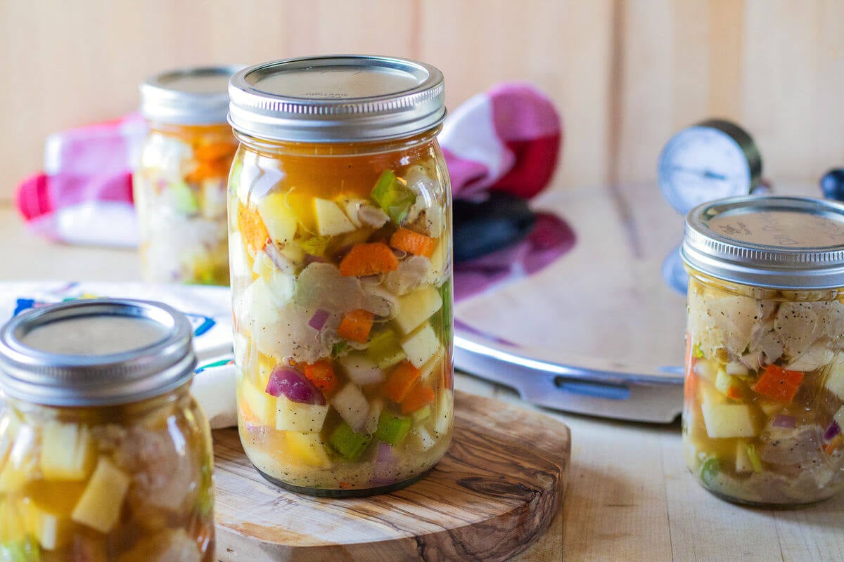 Lidded jars of chicken and vegetables on a wooden counter beside canning equipment.