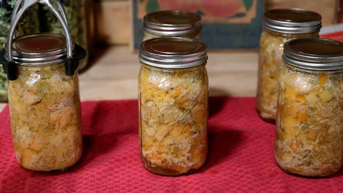 Finished jars of chicken and gravy on a red towel, with a jar lifter holding one jar.