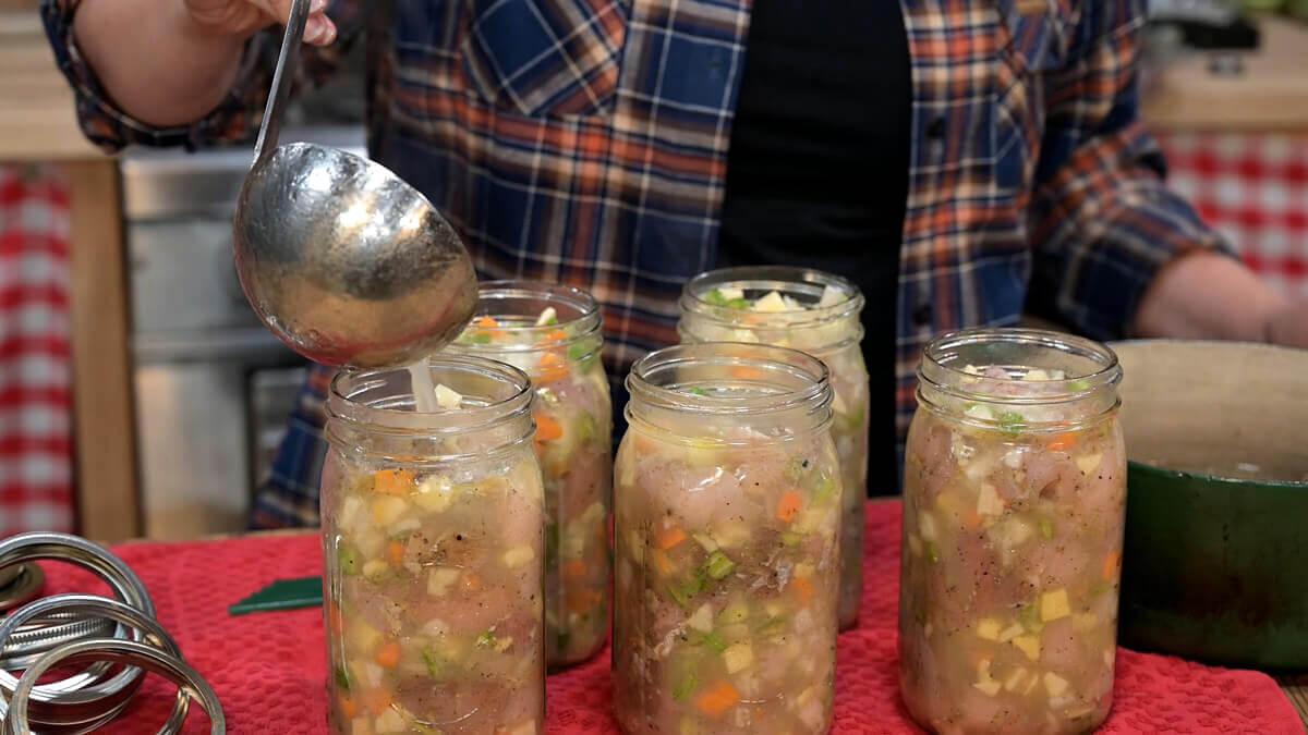 Broth being ladled into jars filled with chicken and vegetables.