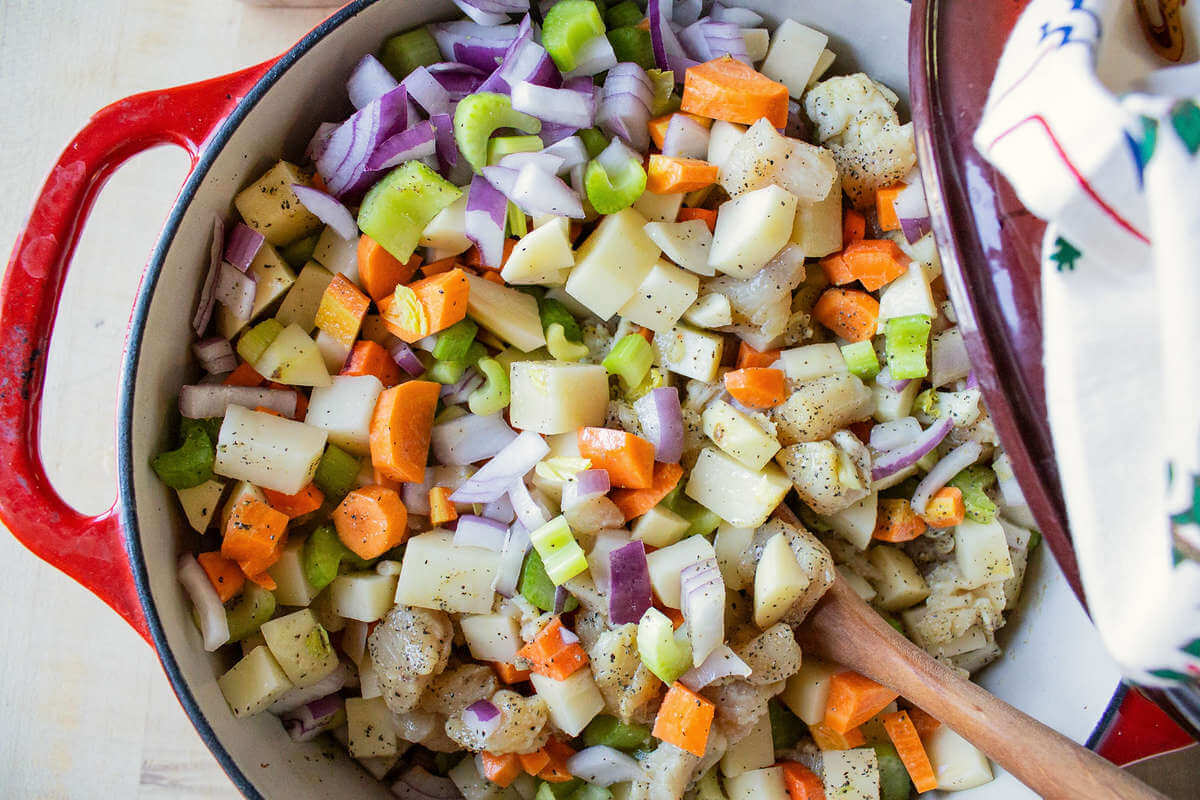 Diced chicken, potatoes, carrots, celery and onions in a red pot with a wooden spoon.