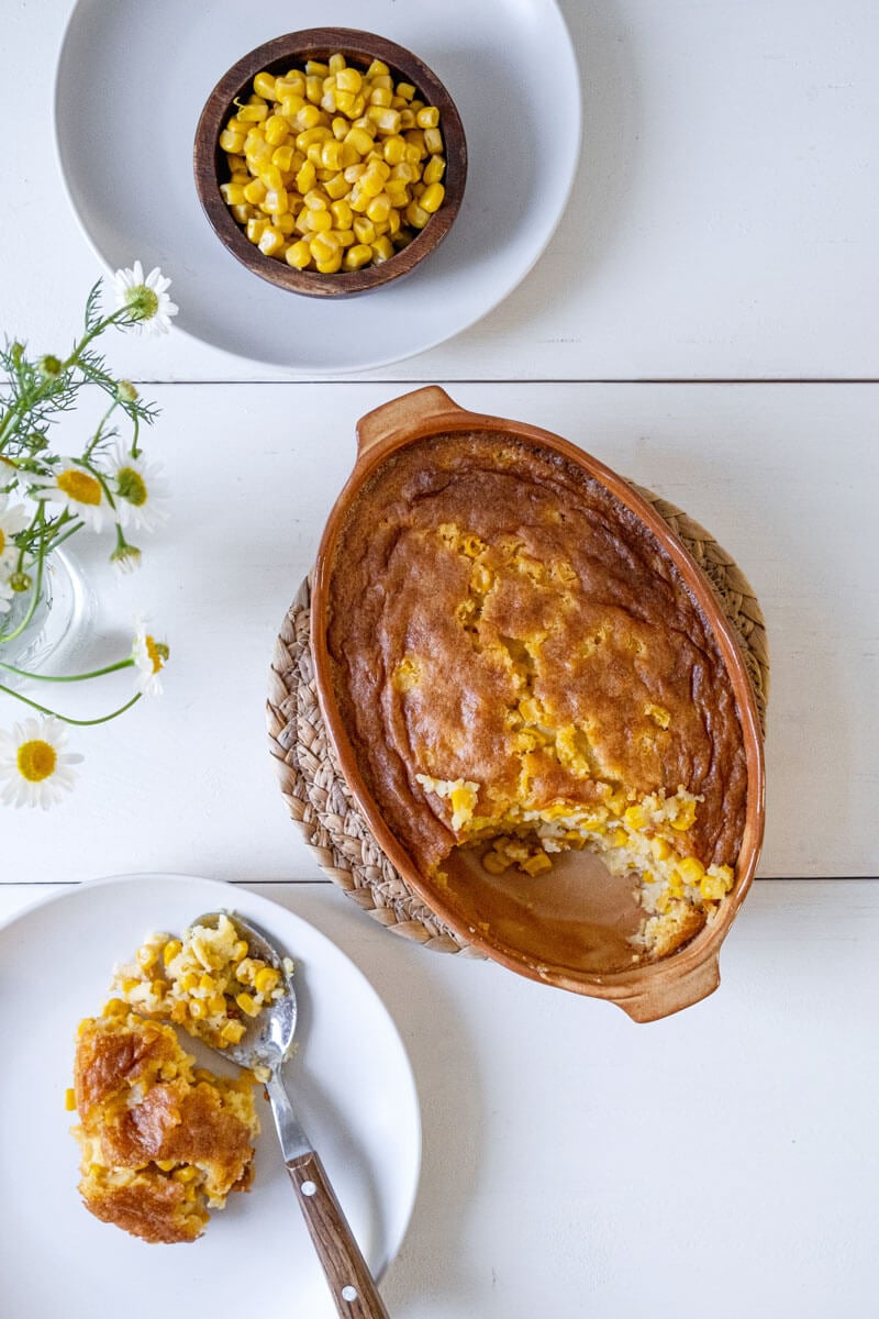 Overhead shot of corn pudding in a casserole dish with some dished up on a white plate.