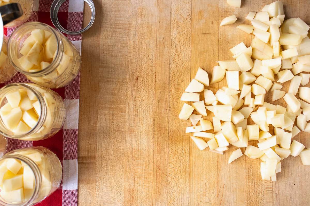 Chopped peeled potatoes on a wooden counter beside open canning jars filled with potato chunks.