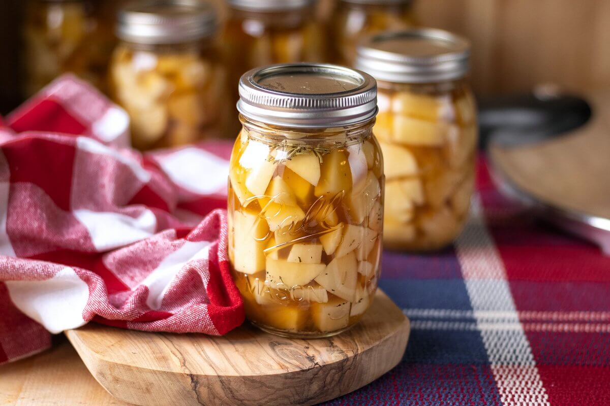 Lidded jar of potato chunks in golden broth on a wooden board, with additional jars behind it.