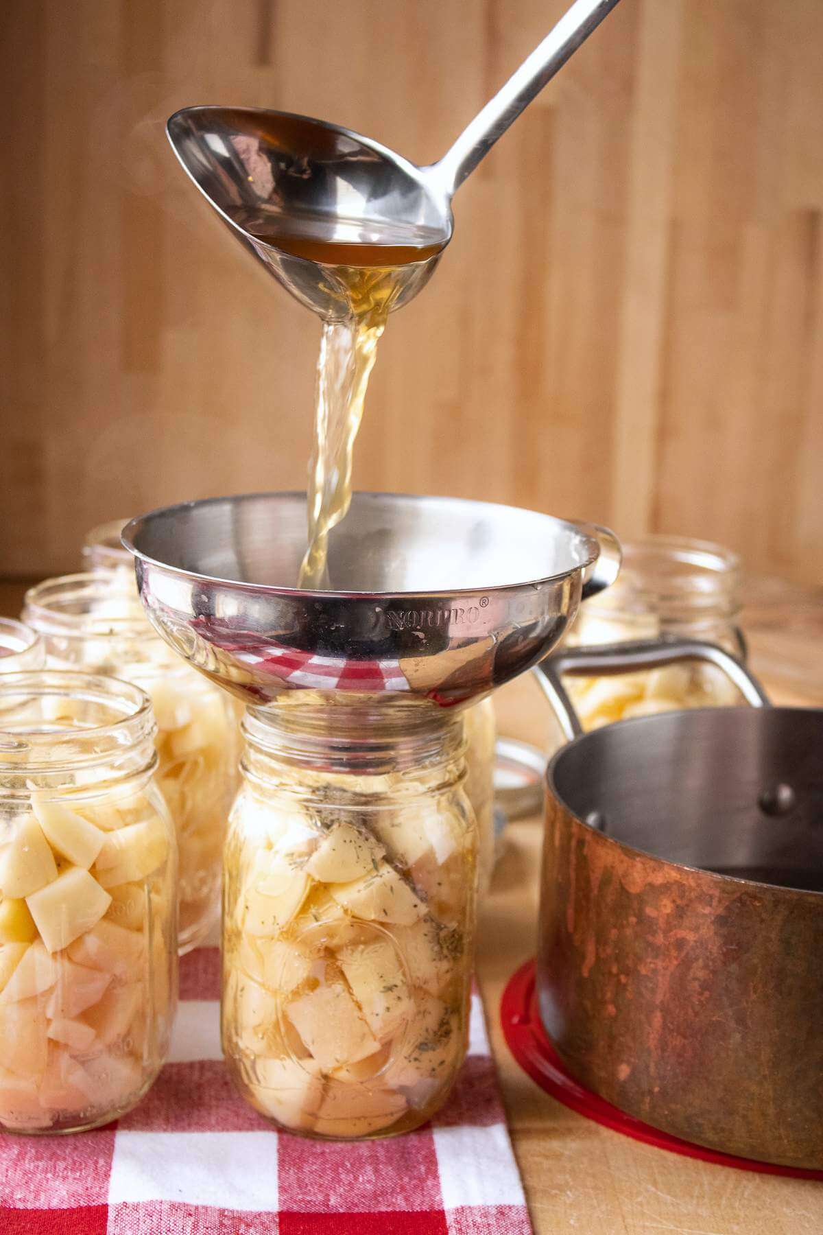 Broth pouring from a ladle through a metal funnel into a jar of potato chunks.