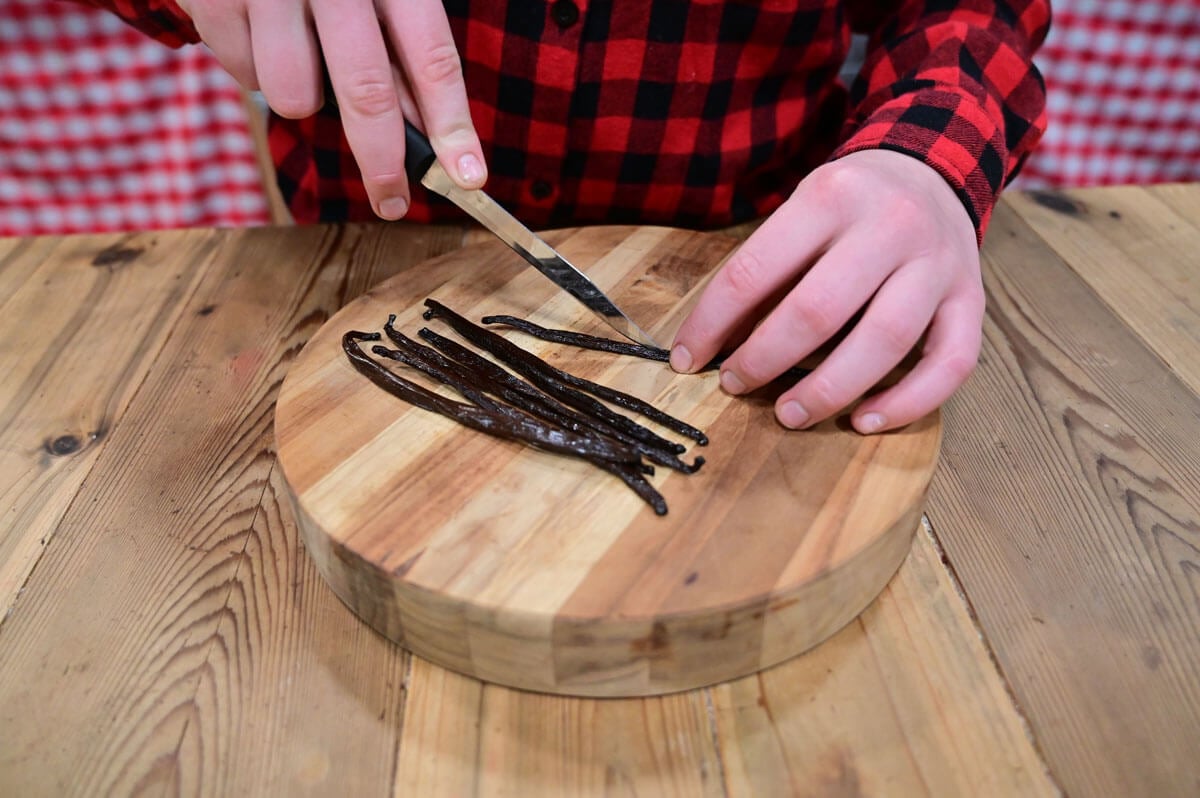 Vanilla beans being sliced in half on a cutting board with a knife.