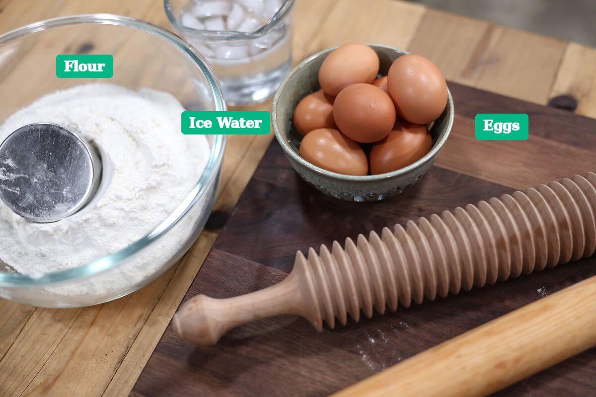 Flour, ice water, and eggs labeled for a homemade egg noodle recipe, with wooden rolling tools.